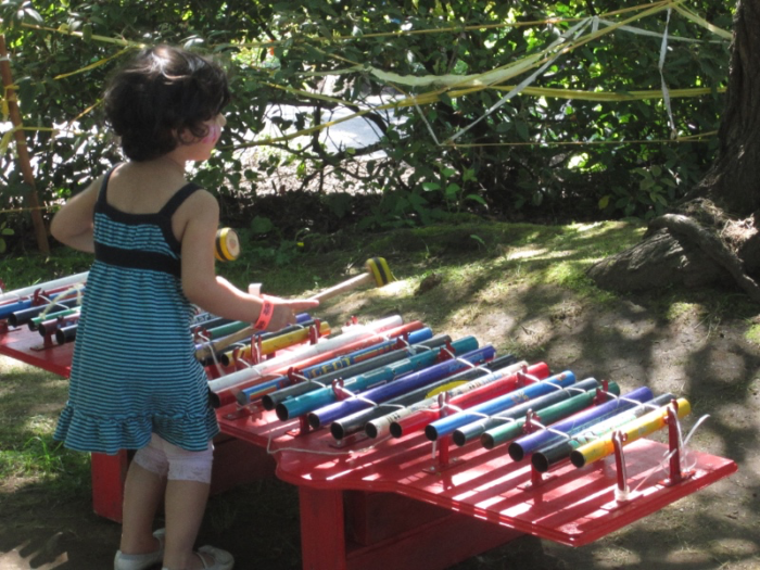 A child plays a multicolored gamelan in a park, using two mallets to strike pipes of differing lengths.