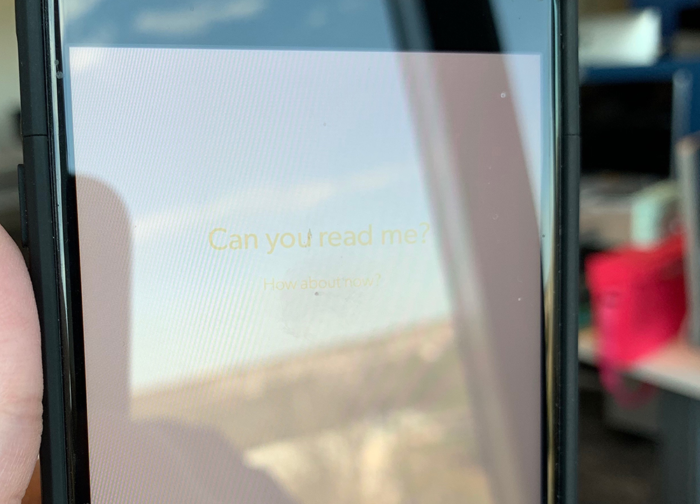 A phone with a heavy glare from a sunny day. There is yellow text on a white background in diminishing sizes, reading "Can you read me?" and smaller "How about now?"