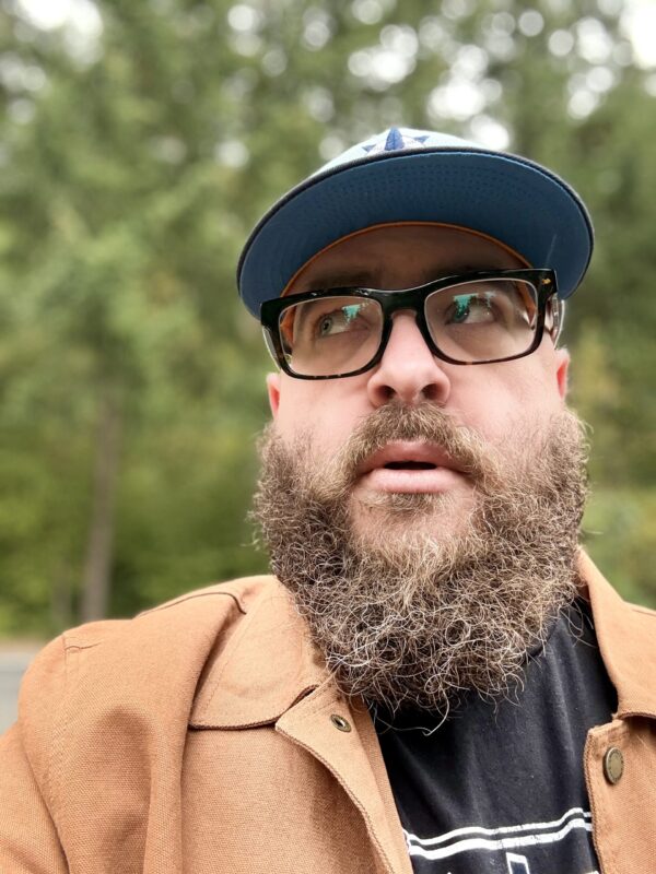 GB glances to his right. He is heavily bearded, wearing thick glasses and a baseball cap. Behind him is the greenery of his home in Washington state.